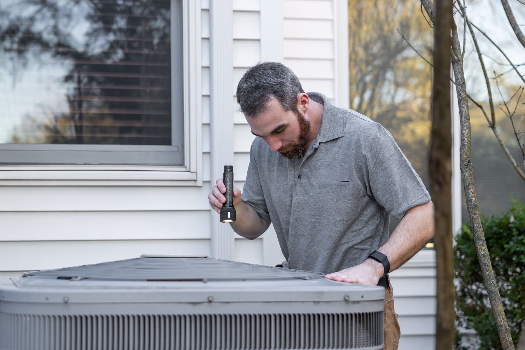 A home inspector in a polo shirt checking the air conditioning unit outside a house with a flashlight